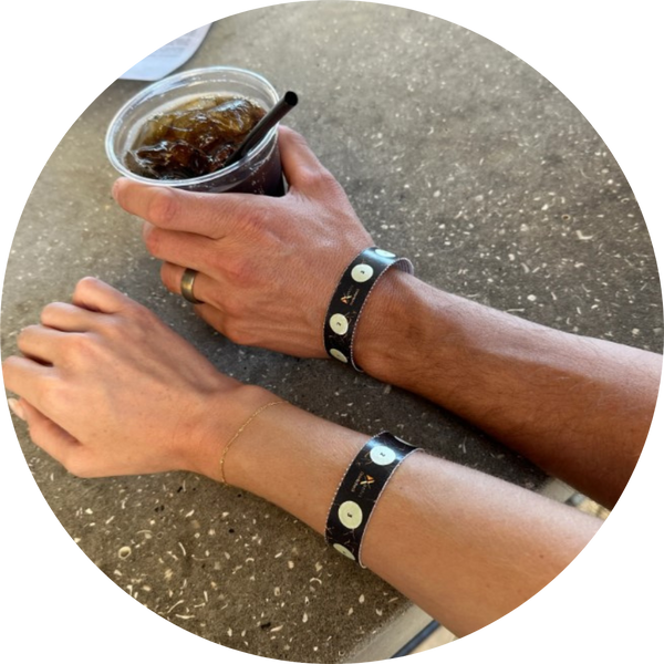 2 hands - one man, one woman - resting on a concrete bar. The man is wearing a wedding band and holding a drink in his hand. The woman is wearing a gold bracelet. Both hands are wearing the Xantus black & gold Drinksafe Wristband.