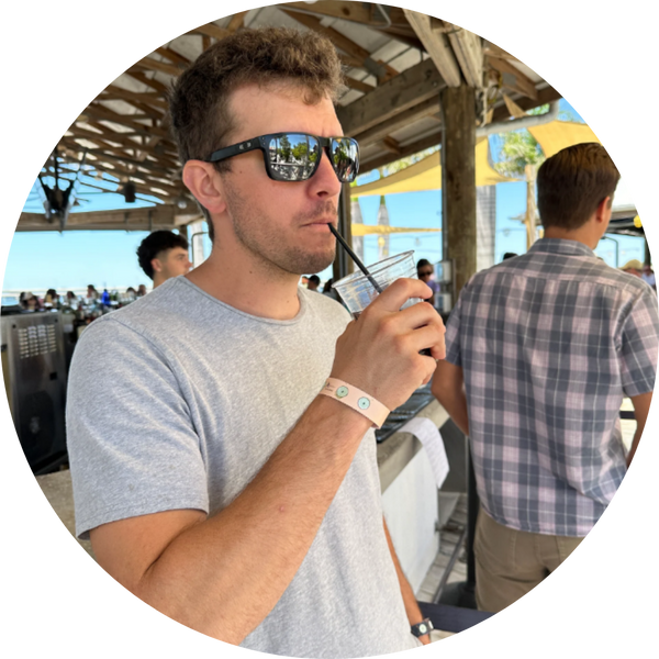 Man with brown hair and mirrored sunglasses at an outdoor bar holding a drink and wearing a sand-colored Xantus Drinksafe Wristband.