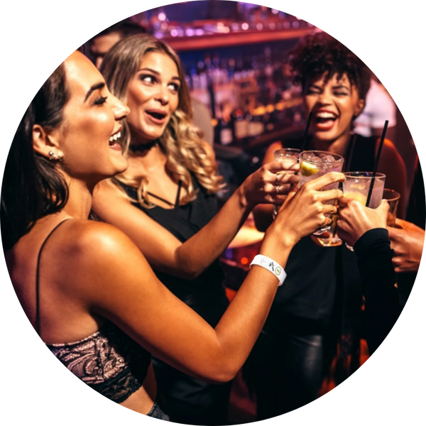 Three women at a bar with drinks in their hands having a good time. One woman is wearing a white Xantus Drinksafe Wristband.